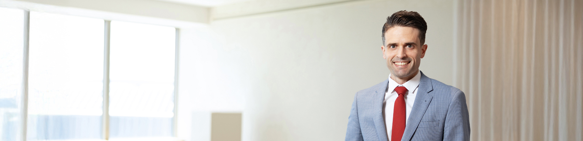 Troy Pilkington, Brunette man in navy suit with red tie leaning against a pole in a white room in front of some windows. A table and chairs and a few green leafy plants are out of focus in the background